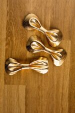 Three gold metallic leather bow ties arranged diagonally on an oak wood surface, displaying the reflective herringbone-patterned leather and handcrafted form.