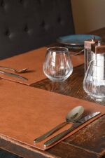 Two tan leather placemats styled on a dark wood dining table with silver cutlery, a stemless wine glass, crystal glassware, and copper salt and pepper mills, with a black tufted leather booth seat in the background.
