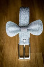 Silver glitter leather bow tie attached to a matching silver glitter belt with a polished metal buckle, photographed vertically on a wooden surface showing the full belt-and-bow-tie combination.