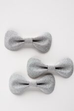 Three silver glitter leather bow ties arranged in a staggered layout on a white background, displaying slight variations in size and showing the consistent glitter texture across multiple pieces.
