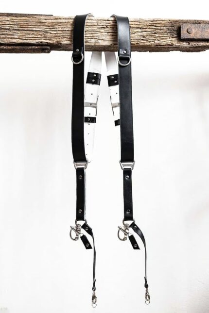 Black GenuineStrap SW Edition leather camera strap with white inner padding, showing D-ring ends hanging over wooden beam