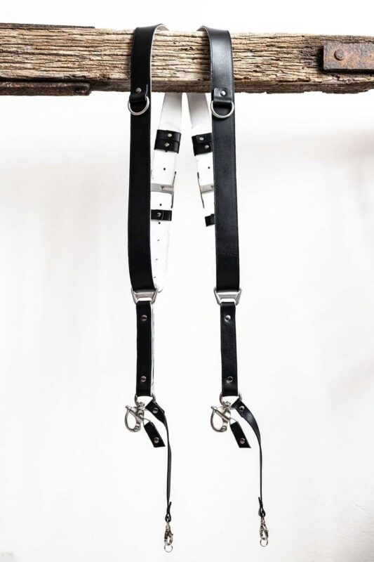 Black GenuineStrap SW Edition leather camera strap with white inner padding, showing D-ring ends hanging over wooden beam