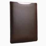 Dark brown leather MacBook sleeve displayed at angle, showing rich leather color, stitched seams, and curved protective shape