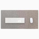 Top-down view of a light gray leather desk mat with stitched edges on a white background, with a white full-size wireless keyboard centered on the left and a white wireless mouse on the right.