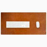 Top-down view of a rich cognac-tan leather desk mat with natural patina and subtle edge stitching, with a white full-size wireless keyboard and white wireless mouse placed on it, on a white background.