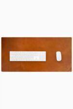 Top-down view of a rich cognac-tan leather desk mat with natural patina and subtle edge stitching, with a white full-size wireless keyboard and white wireless mouse placed on it, on a white background.