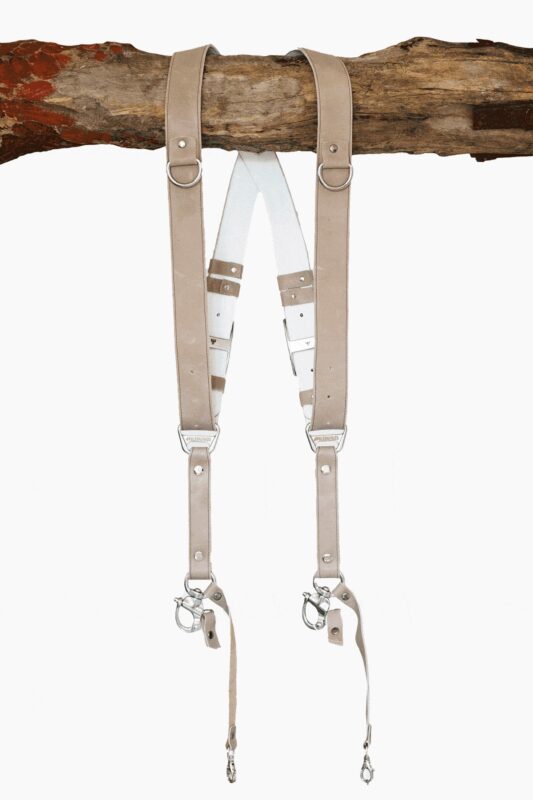 Sand-colored vintage leather dual camera harness with silver hardware, hanging from a rustic wooden branch against a white background, showing the full front view with two shoulder straps, a white leather back panel, branded "GENUINESTRAP" metal connectors, and snap hook attachments at each end.